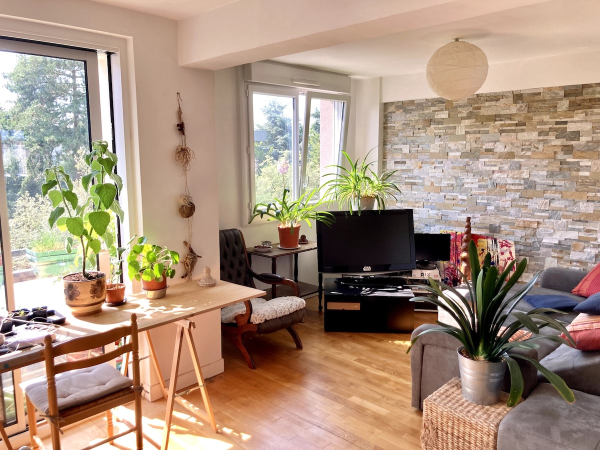 A bright living space is shown, featuring large windows that allow natural light to enter. A comfortable sofa is positioned near a stone accent wall, adorned with various indoor plants. A wooden table and chairs are set beside the window, creating a cozy dining area.