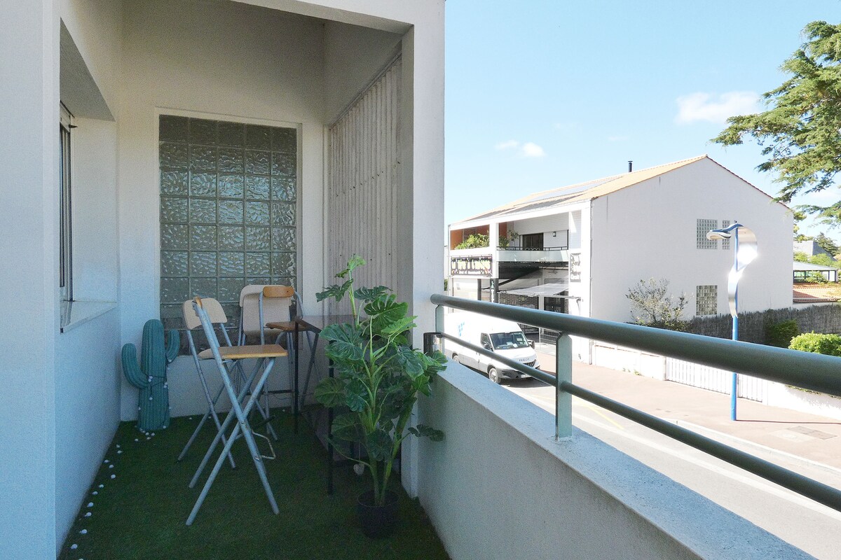 A small balcony is visible, featuring two chairs and a table positioned on a green lawn-like surface. A potted plant adds a touch of greenery. The railing provides a view of the surrounding area, including nearby buildings and clear blue skies.