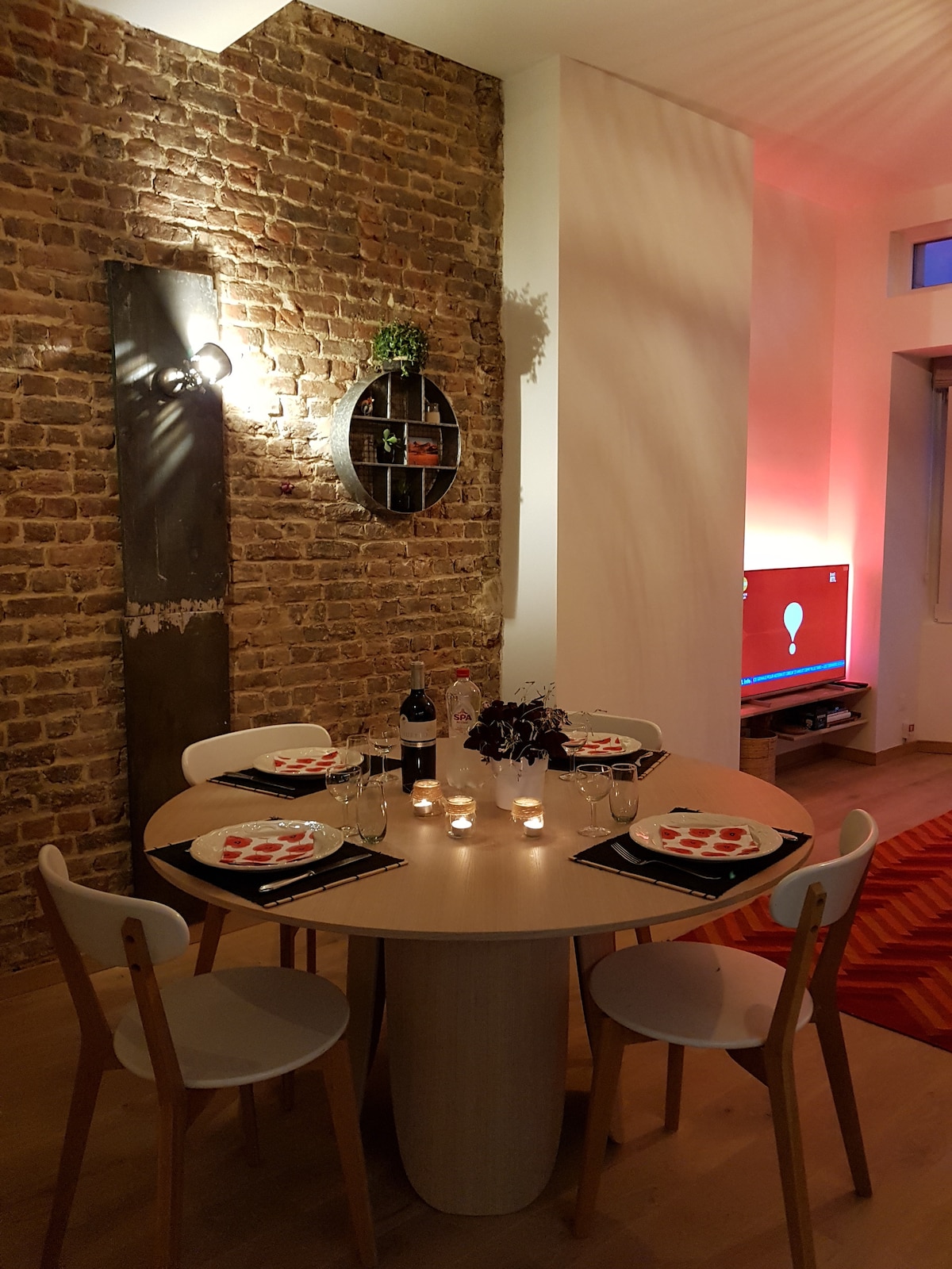 A dining area features a round table set for four, adorned with plates, cutlery, and candles. Exposed brick walls enhance the space, while a small shelf displays decorative items. Subtle lighting adds warmth, with a television visible in the background.