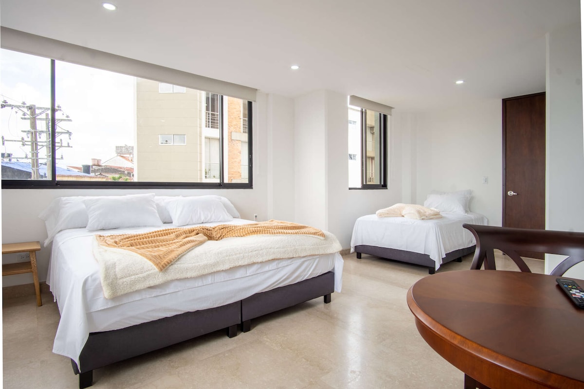 The room features two spacious beds dressed in white linens, accented with soft throws. Large windows bring in natural light, showcasing concrete and city views. A round wooden table is positioned near one bed, complementing the overall minimalist aesthetic.