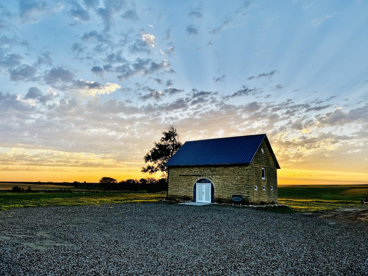 A historic stone barn is positioned against a stunning sunset, with rays of light illuminating the sky. The surrounding landscape features open fields under a colorful horizon, while a gravel pathway leads to the barn's entrance.