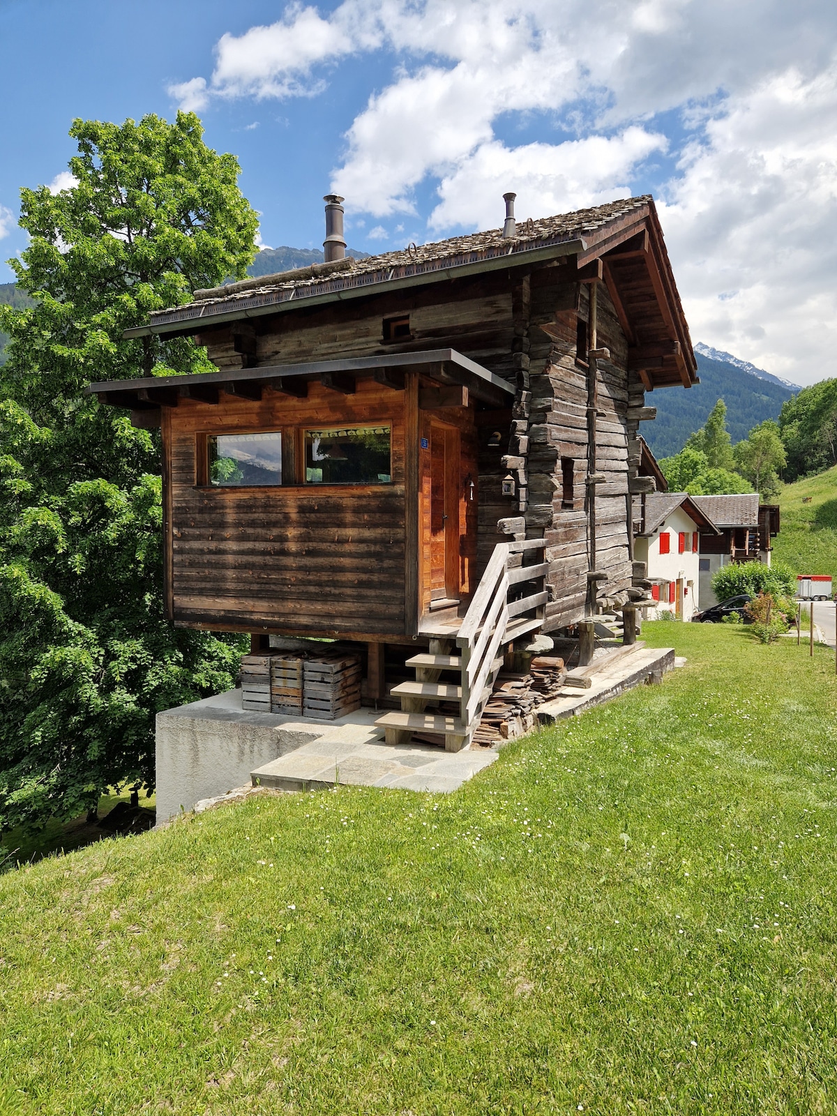 A traditional wooden structure is set on a grassy area, surrounded by trees and mountains. The exterior features rustic wooden panels and a small staircase leading to the entrance. Nearby, a second building can be seen with a contrasting color scheme.