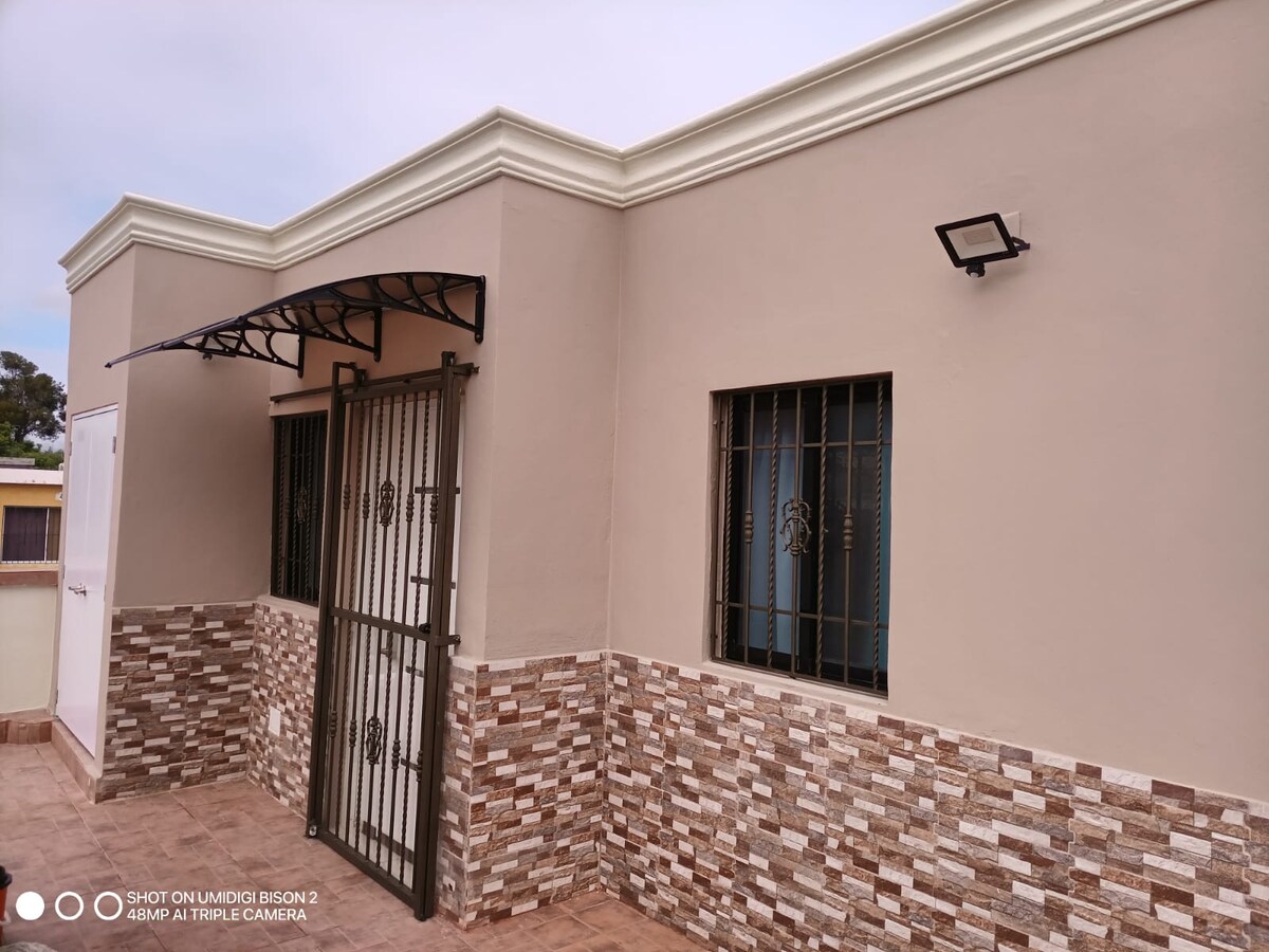 The exterior of the building is shown, featuring a smooth beige wall complemented by a textured stone base. A decorative wrought iron gate stands prominently, while two windows with metal grilles are positioned symmetrically. An awning provides shade above the entrance.
