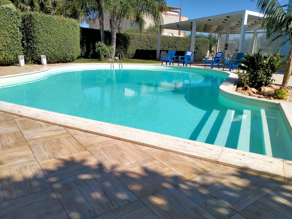 A serene pool area is framed by green hedges and palm trees. Bright blue lounge chairs are positioned nearby, inviting relaxation. The clear water reflects sunlight, enhancing the tranquil setting. A shaded pergola is visible in the background, providing additional seating options.