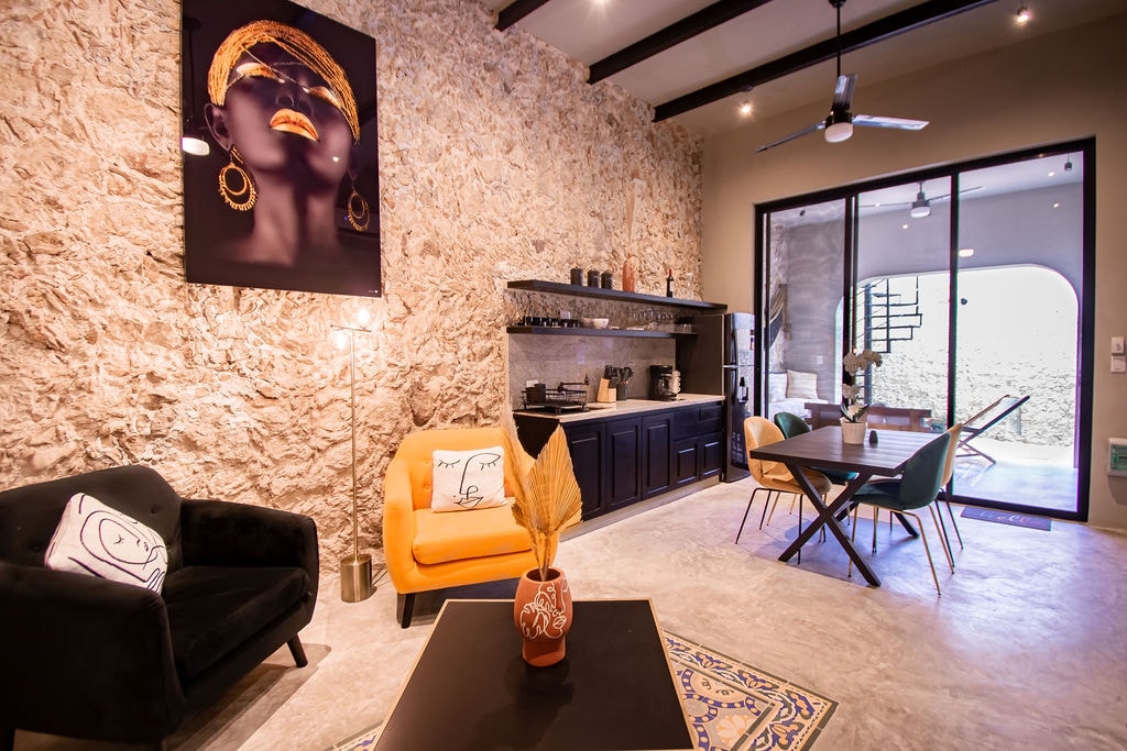 An inviting living area features a textured stone wall and contrasting dark cabinetry. A modern black sofa and a vibrant orange chair are positioned around a sleek coffee table. Natural light filters through glass doors, leading to an outdoor space, while a dining table awaits nearby.