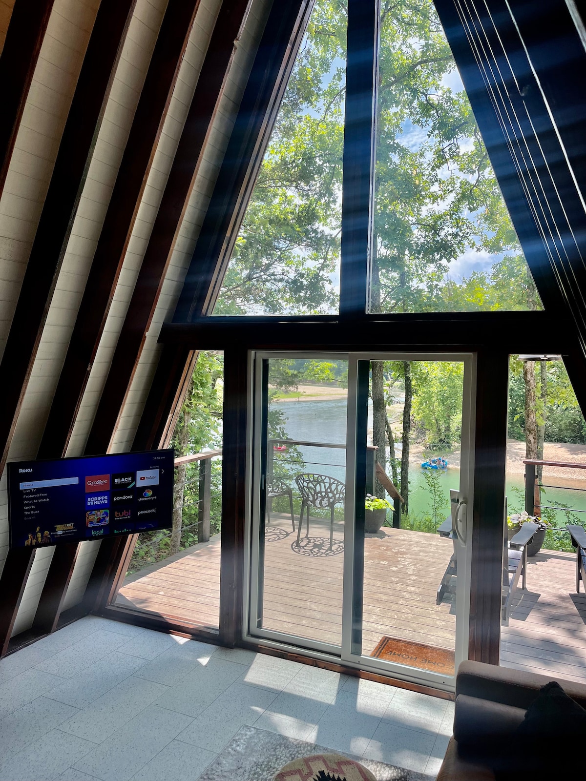 Natural light floods a modern a-frame cabin's interior, highlighting the sloped wooden beams. A Roku TV is mounted on the wall, while large glass doors offer views of the Illinois River. A small outdoor deck features seating and scenic water access surrounded by trees.