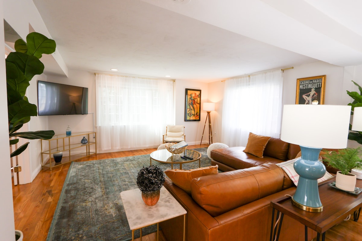 The spacious living room features a leather sectional sofa and a large coffee table surrounded by greenery. A 55-inch TV is mounted on the wall opposite a bright window, which is adorned with sheer curtains, offering natural light, while a floor lamp provides additional illumination.