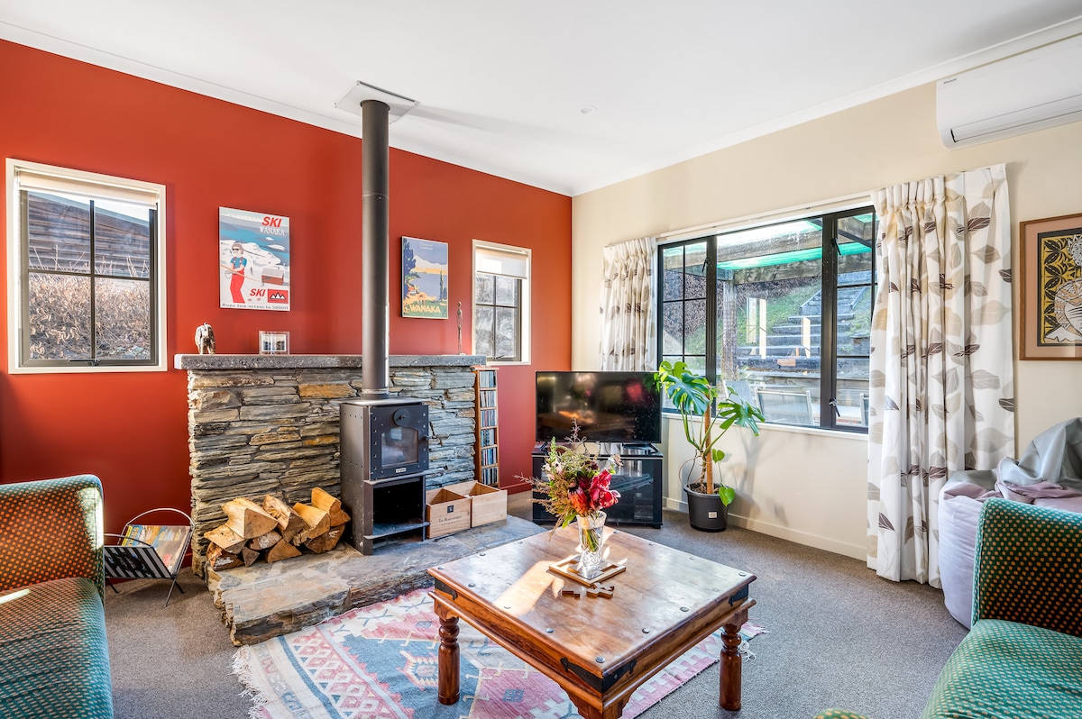A spacious lounge area is highlighted by a rustic stone fireplace and a log burner. Large windows allow natural light to fill the room, complemented by colorful wall art and a decorative table at the center, featuring a floral arrangement.