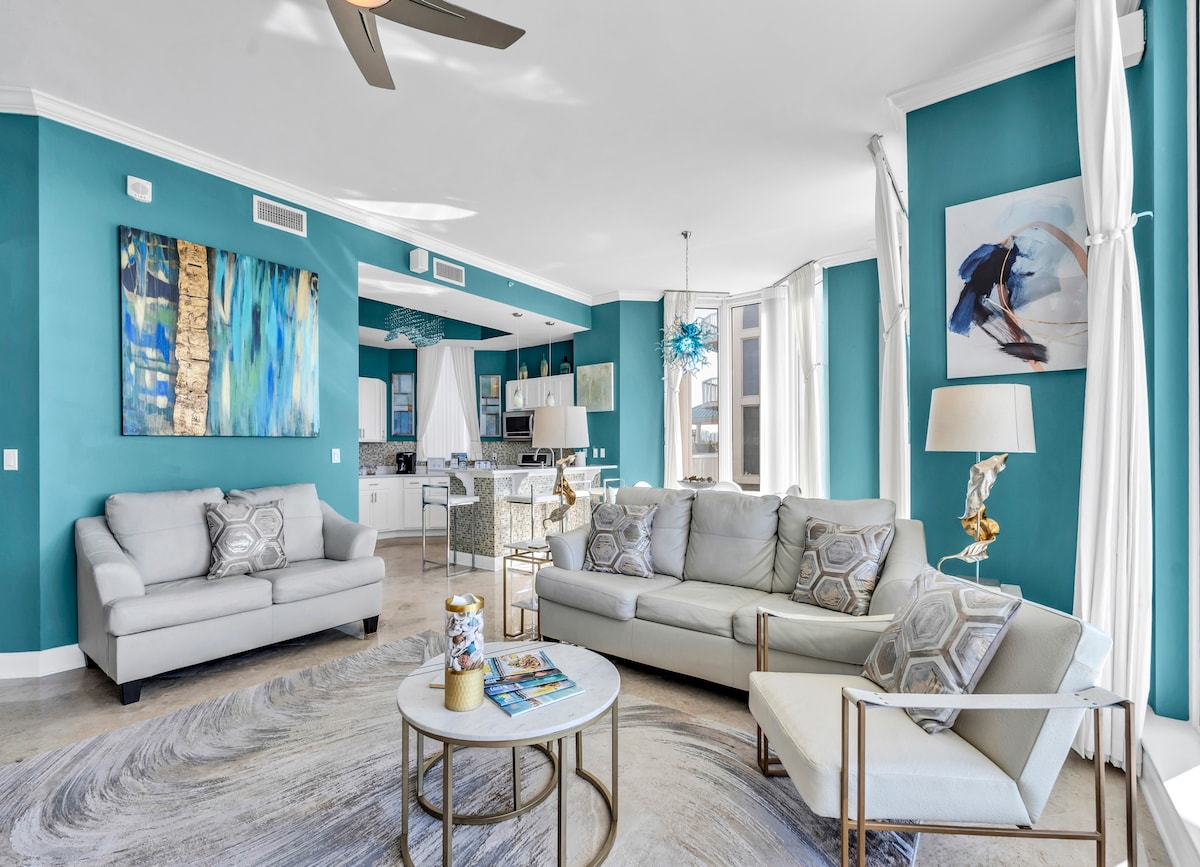 A bright living area is highlighted by teal walls and large windows allowing natural light. Two sofas are arranged around a round coffee table, complemented by decorative pillows. Artwork adorns the walls, and a modern ceiling fan is visible above.