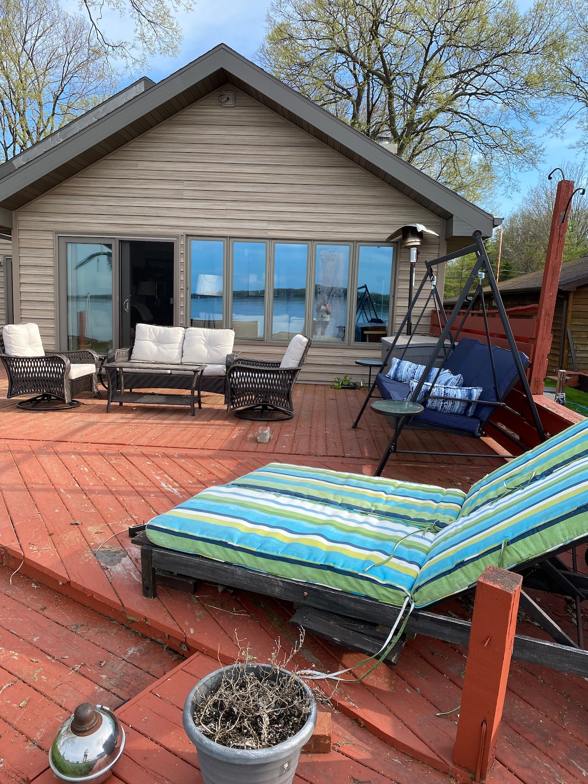 An outdoor deck features a variety of seating options, including a swing and lounge chair with striped cushions. Large windows in the background provide views of the lake, while the deck's wooden surface adds a warm touch to the space.