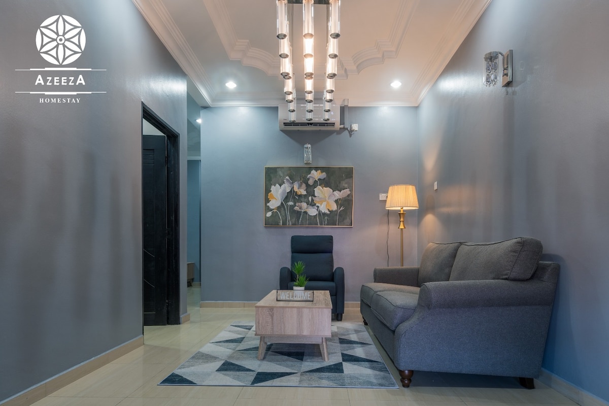 A modern living room is presented with a soft grey sofa and a small armchair. A wooden coffee table sits centrally on a light rug. The walls are painted in a calm blue tone, accompanied by an artwork that adds a touch of nature to the space. Ambient lighting is provided by a unique ceiling fixture.