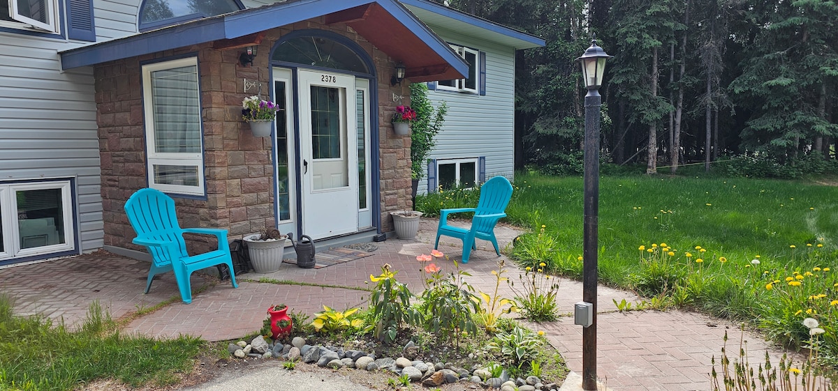 An inviting entrance showcases a covered porch with two blue chairs and potted flowers. A winding stone pathway leads through a garden area with greenery and wildflowers. Nearby, a lamp post provides subtle illumination, with trees visible in the background.