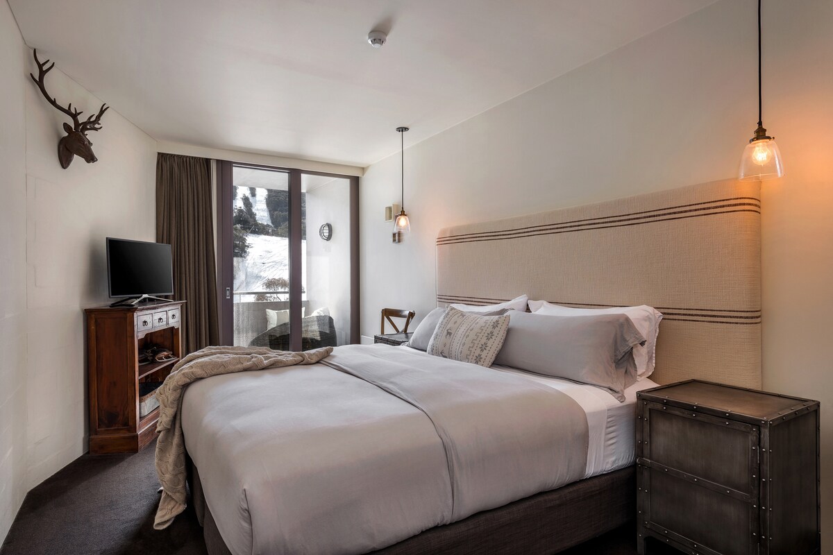 A cozy bedroom features a spacious bed with soft, neutral bedding. Light wood furniture is complemented by a wooden chest and a television. Soft lighting is provided by hanging fixtures, while large windows offer a view of the surrounding landscape.