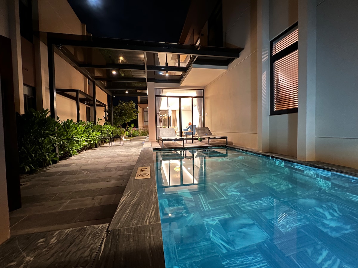 An evening view of a tranquil pool area is captured, showcasing a sleek swimming pool with vibrant blue water. Comfortable lounge chairs are positioned beside the pool, framed by lush greenery and well-lit pathways extending toward the villa's entrance.
