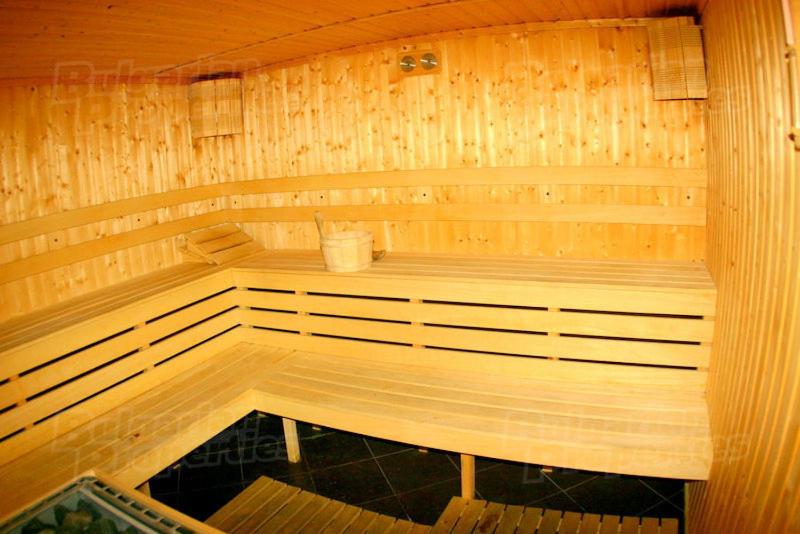 A wooden sauna interior is depicted, featuring multiple benches arranged in a L-shape. Soft lighting from wall-mounted fixtures enhances the warm ambiance. A wooden bucket is placed on a bench, while the textured walls and floor create a natural and calming environment.
