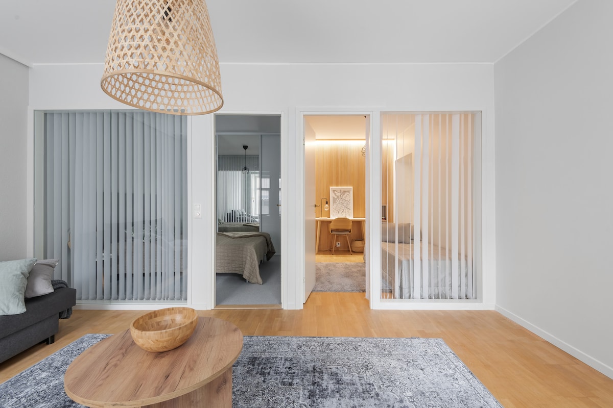 A spacious living area is highlighted by natural wooden flooring and a large, round coffee table. Two bedrooms are visible through sheer curtains, creating a sense of openness. A woven pendant lamp adds a touch of warmth to the contemporary design.