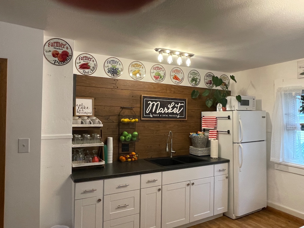 A modern kitchen features a wooden accent wall adorned with decorative plates. A dark countertop complements the white cabinetry, while a double sink is positioned under a wall-mounted sign that reads 'Market.' Fresh fruits are displayed in baskets, enhancing the inviting atmosphere.