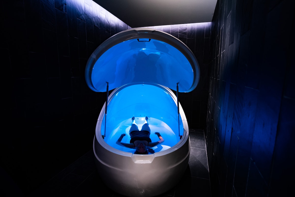 A sensory deprivation float tank is illuminated by soft blue lighting. The sleek, rounded design is set against dark tiled walls, creating a calm and tranquil environment. Water within the tank reflects the gentle glow, enhancing the serene atmosphere.