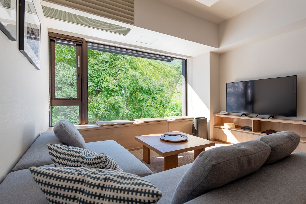 A lounge area is highlighted by a large window offering views of lush greenery. A comfortable grey sofa with patterned cushions faces a wooden coffee table, complemented by a television on a media unit. Natural light fills the spacious room, enhancing its inviting ambiance.