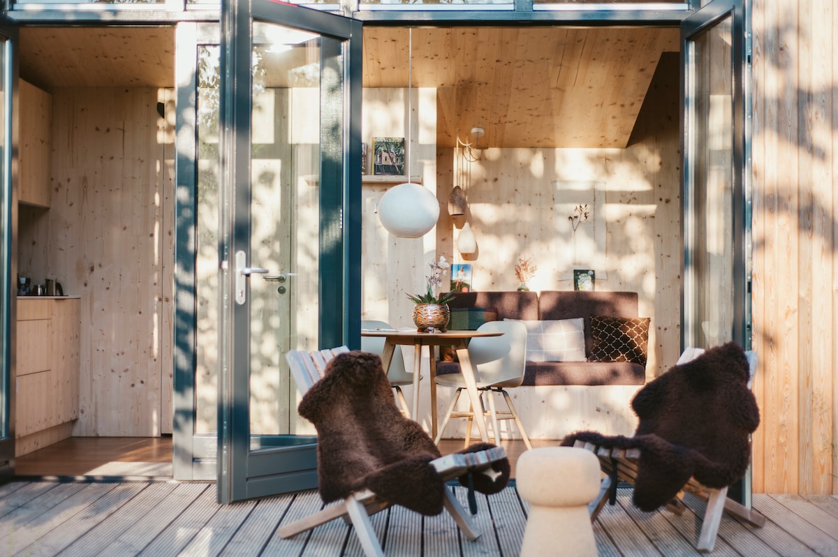An inviting outdoor seating area features two comfortable chairs with soft coverings, positioned on a wooden deck. Large glass doors open to a light-filled interior, where a small dining table and cozy seating can be seen, complemented by warm wood tones and natural textures.