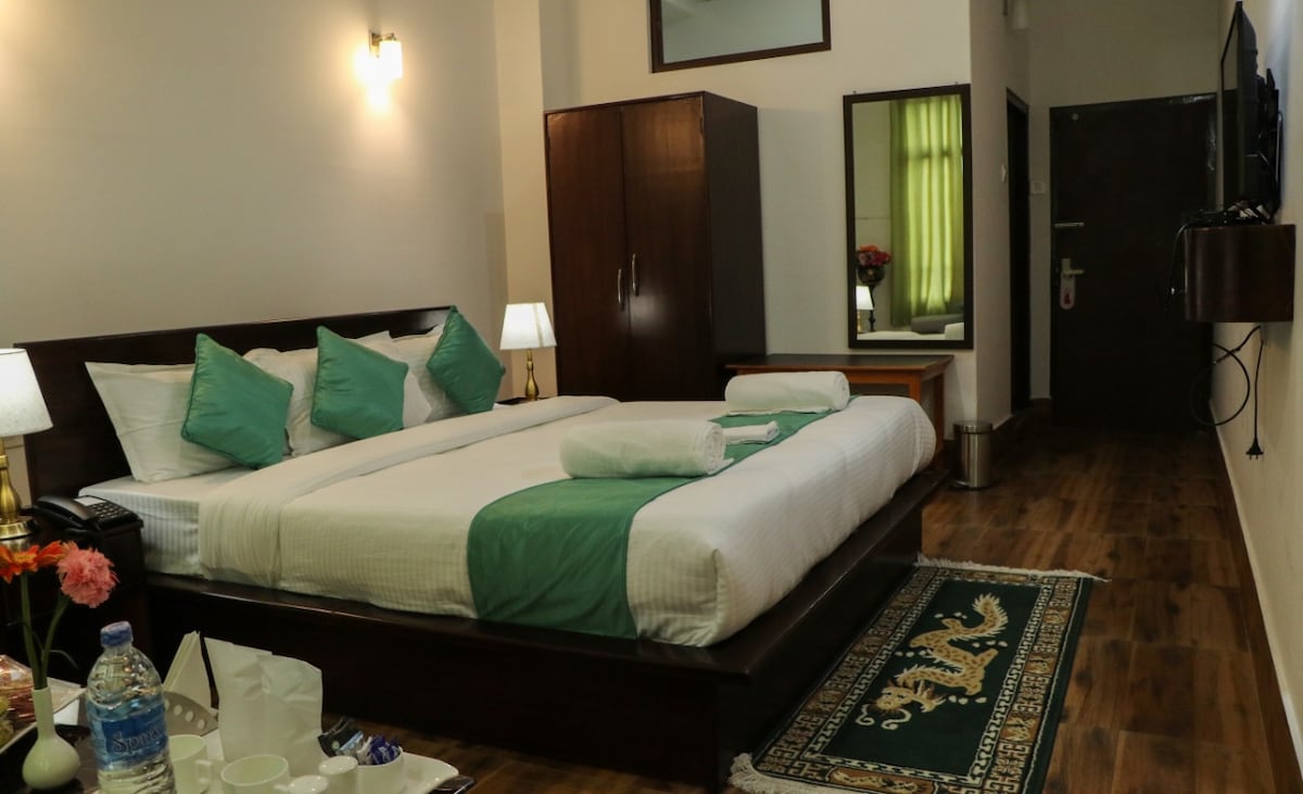 A well-appointed room features a spacious bed with a white comforter and green accents. Two bedside lamps provide soft lighting, while a mirrored cabinet reflects the green curtains. A small seating area with a wooden table and a patterned rug completes the warm ambiance.