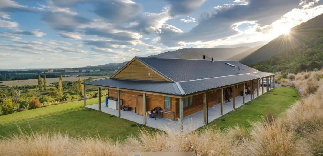 The single-story house is set against a backdrop of rolling hills and expansive skies. A spacious porch wraps around the exterior, complemented by lush grass and native plants. Sunlight casts a warm glow over the structure, enhancing its natural wood finishes and inviting design.
