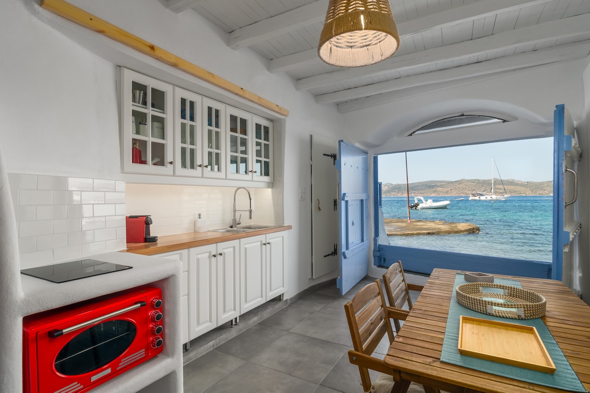 A light-filled kitchen features modern appliances, including a bright red stove. A wooden dining table and chairs are positioned near an open door that offers a view of the sea and anchored boats. Cabinets line the wall, providing ample storage.