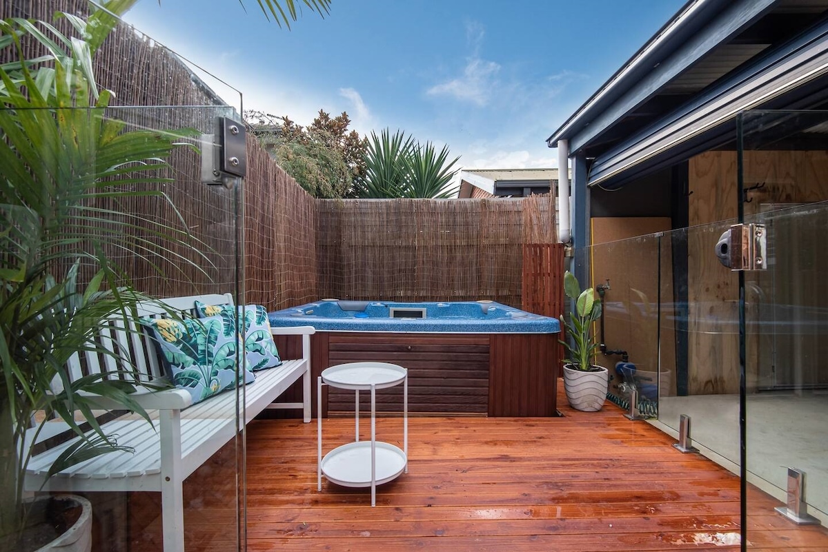 A private outdoor area showcases a jacuzzi spa surrounded by tall greenery for added privacy. A comfortable white bench with decorative cushions is positioned next to a small round table, creating a relaxing space on polished wooden decking.