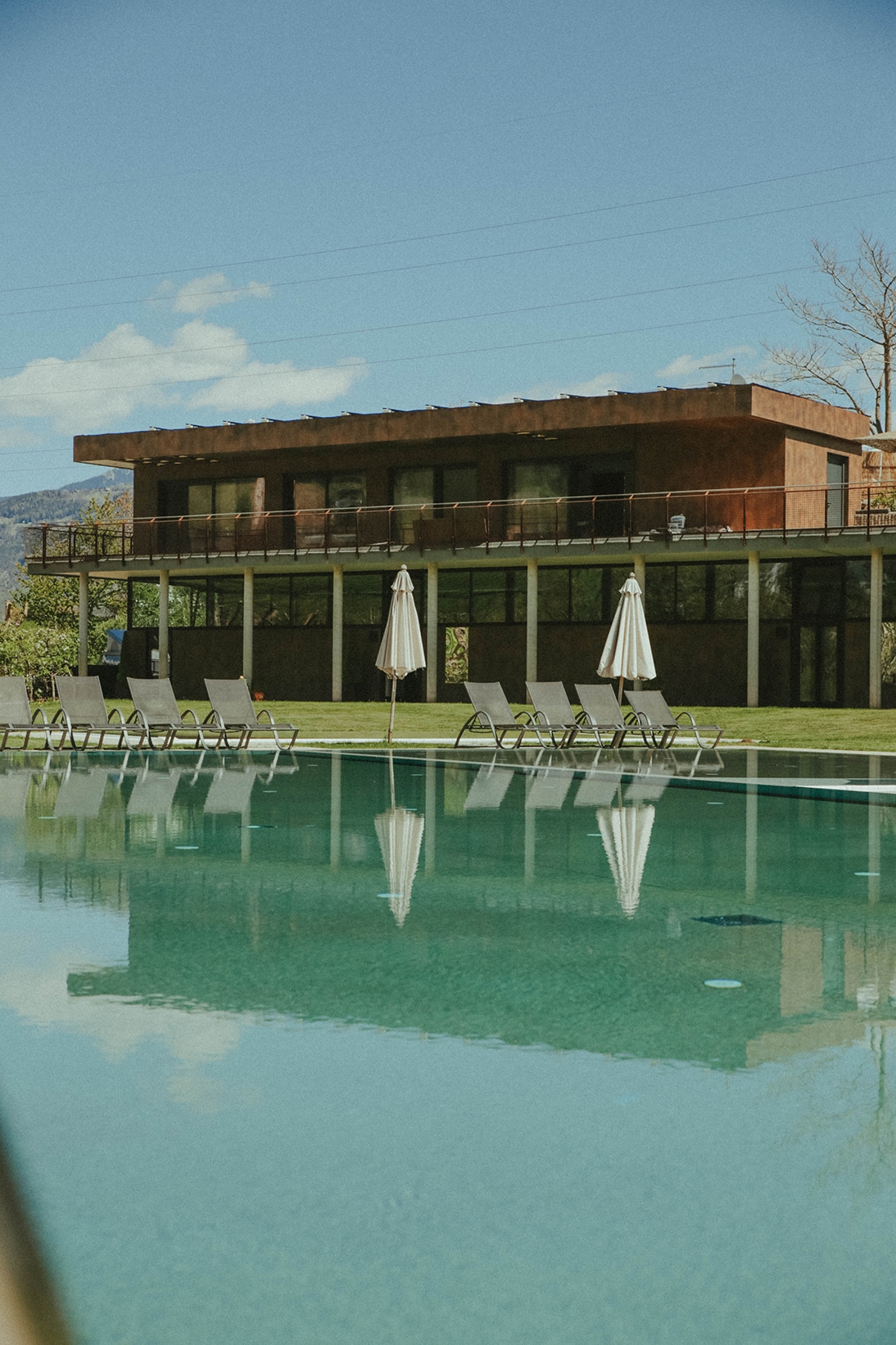 A modern two-story building is reflected in a serene swimming pool, surrounded by a well-maintained lawn. Several lounge chairs are arranged around the poolside, with umbrellas providing shade. The structure features large windows, enhancing the connection to the outdoor landscape.