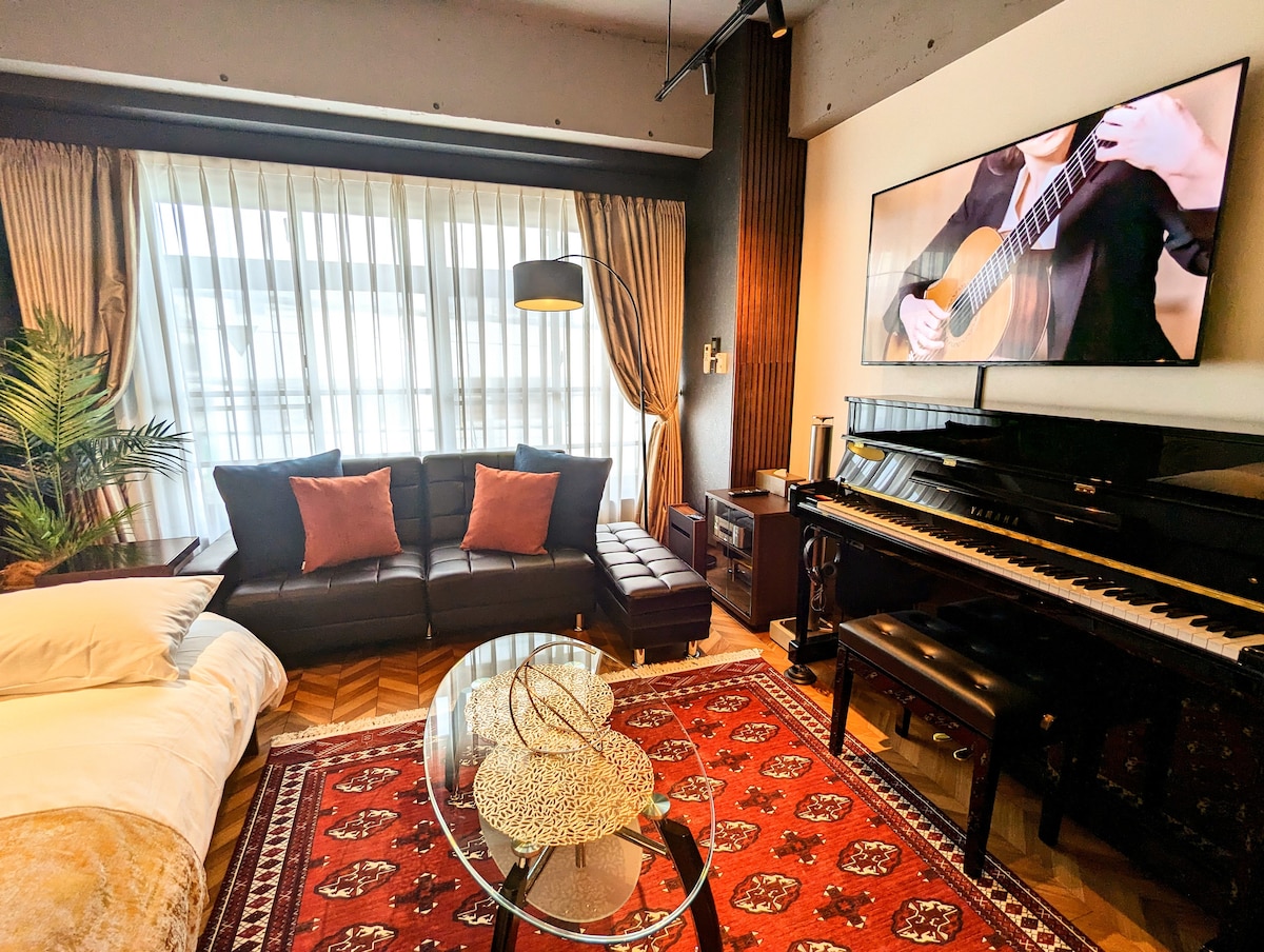 A spacious living area features a dark leather sofa with decorative pillows positioned around a glass coffee table. A Yamaha upright piano stands nearby, while a large television displays a performance. Natural light filters through sheer curtains, enhancing the warm ambiance.