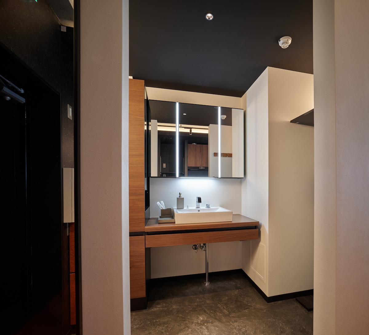 A clean and modern bathroom is showcased, featuring a sleek vanity with a sink and mirror. Minimalistic light fixtures provide a soft glow above the counter. Dark flooring complements the light wood cabinetry and white walls, creating a balanced aesthetic.