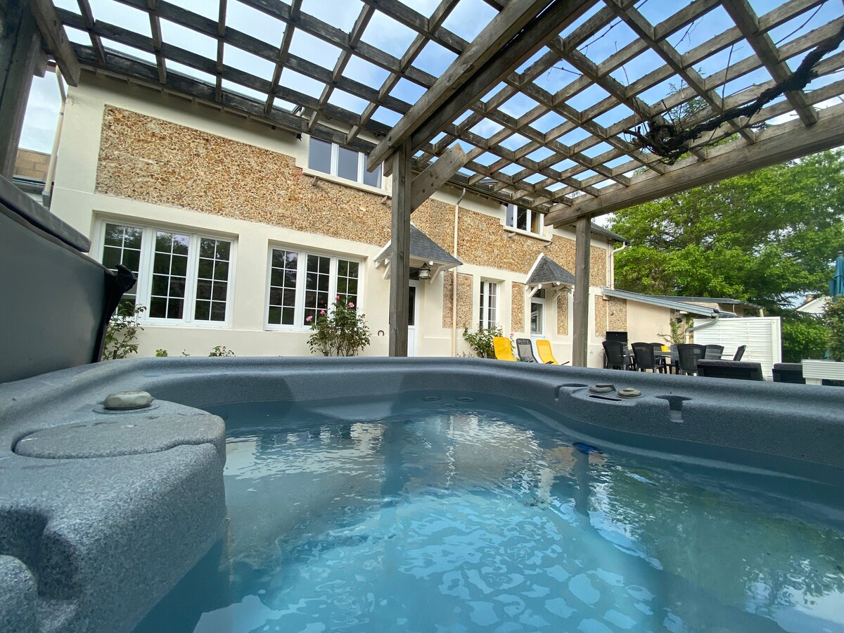A private outdoor jacuzzi is placed under a wooden pergola, offering a view of the house's rustic stone facade. The surrounding area features comfortable seating and bright yellow lounge chairs that invite relaxation. Large windows of the house allow natural light to illuminate the space.
