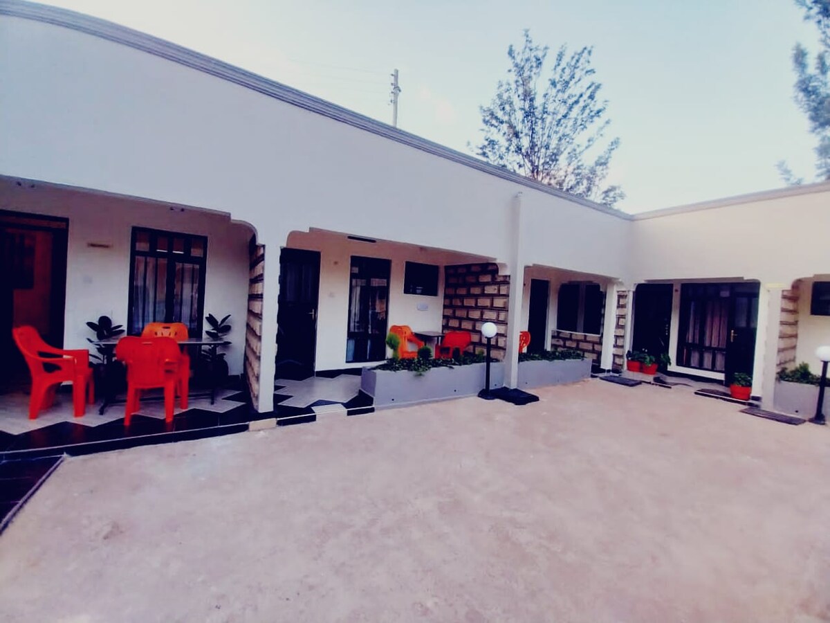 A courtyard surrounded by several rooms features vibrant orange chairs set around small tables. Potted plants are placed along the walls, adding greenery. The structure has a minimalist design, with large windows allowing natural light to enter the rooms.