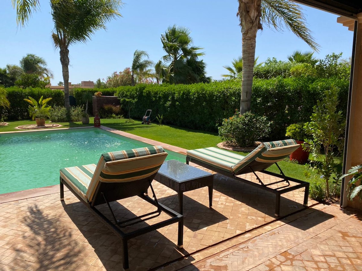 Two lounge chairs with striped cushions are positioned beside a sparkling pool. Lush greenery surrounds the area, with tall palm trees in the background, creating a serene outdoor space ideal for relaxation.