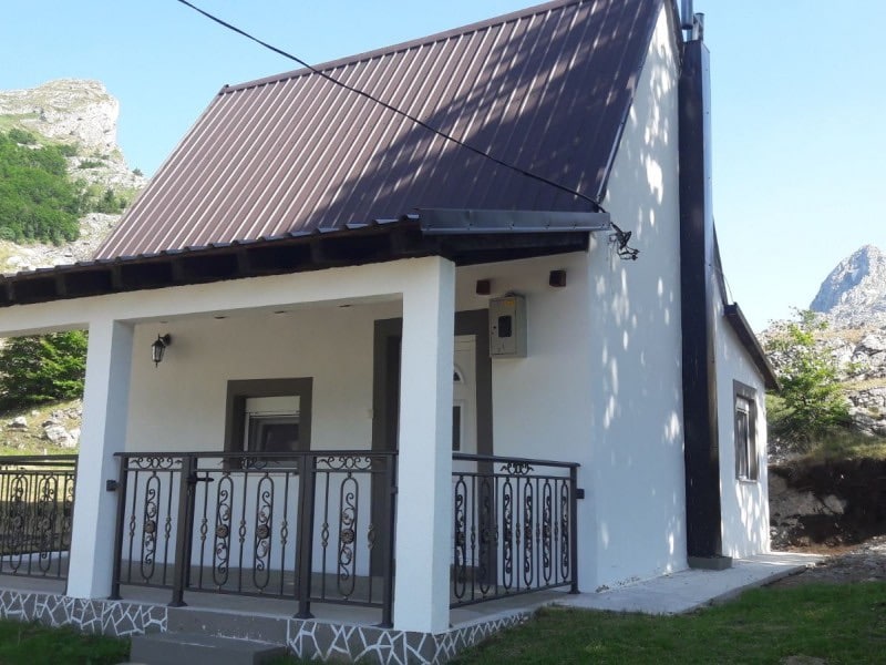The exterior of a charming cabin is presented, featuring a sloped roof with a dark metal finish. A covered porch with a decorative railing offers space for relaxation, while the surrounding nature includes rocky hills and greenery.