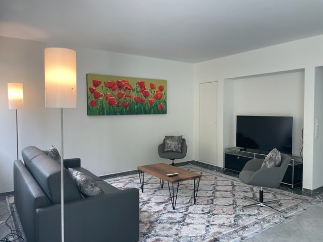 A spacious living area is highlighted by a large abstract painting featuring red tulips. Modern furniture includes a dark sofa, two accent chairs, and a coffee table. A flat-screen TV is positioned on a low media console, with soft lighting provided by sleek floor lamps.