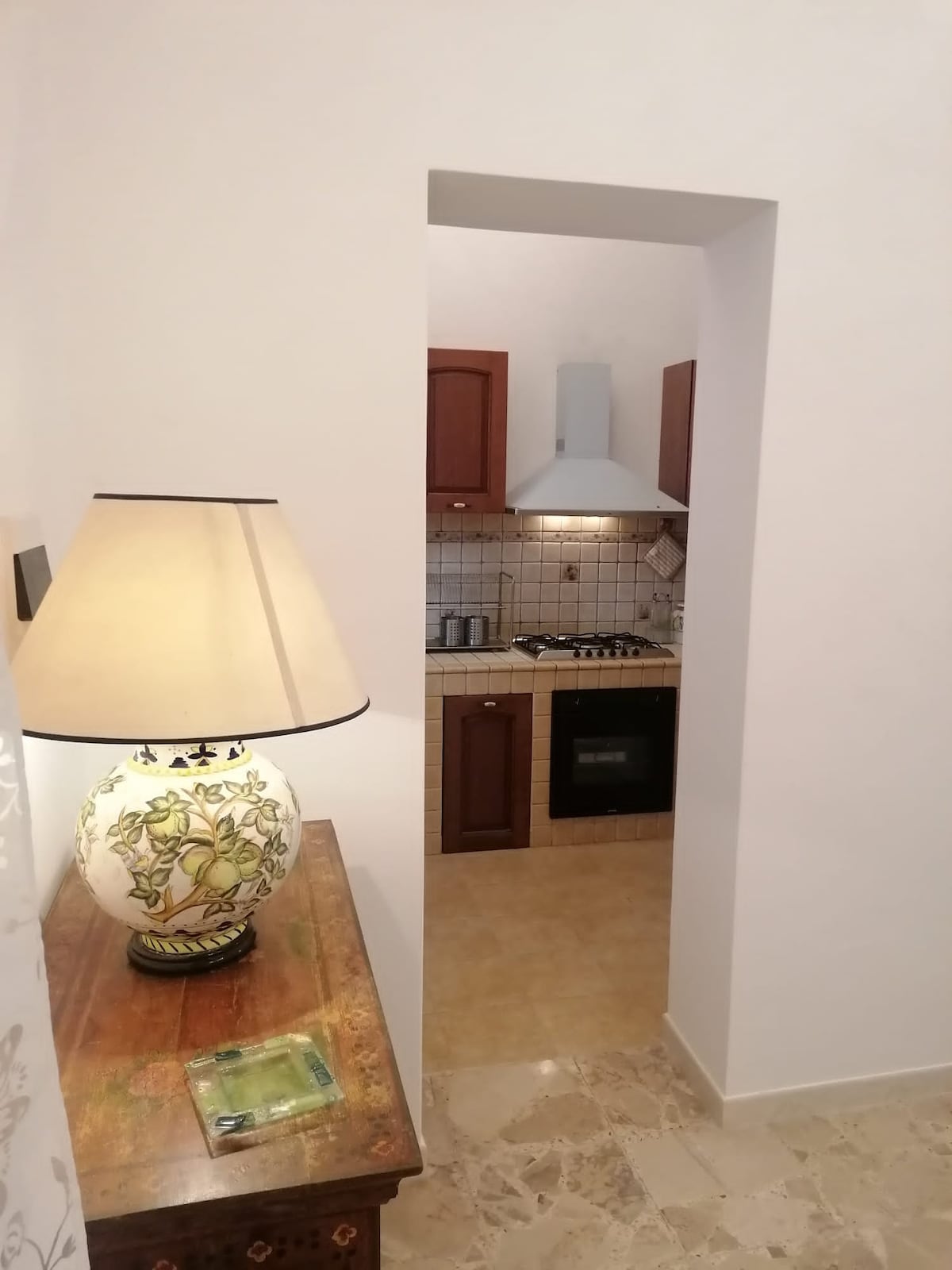 A well-lit corner showcases a decorative lamp atop a wooden table, drawing attention to the soft tones of the surrounding walls. In the background, a compact kitchen is visible, featuring wooden cabinetry, a stove, and essential cooking appliances.