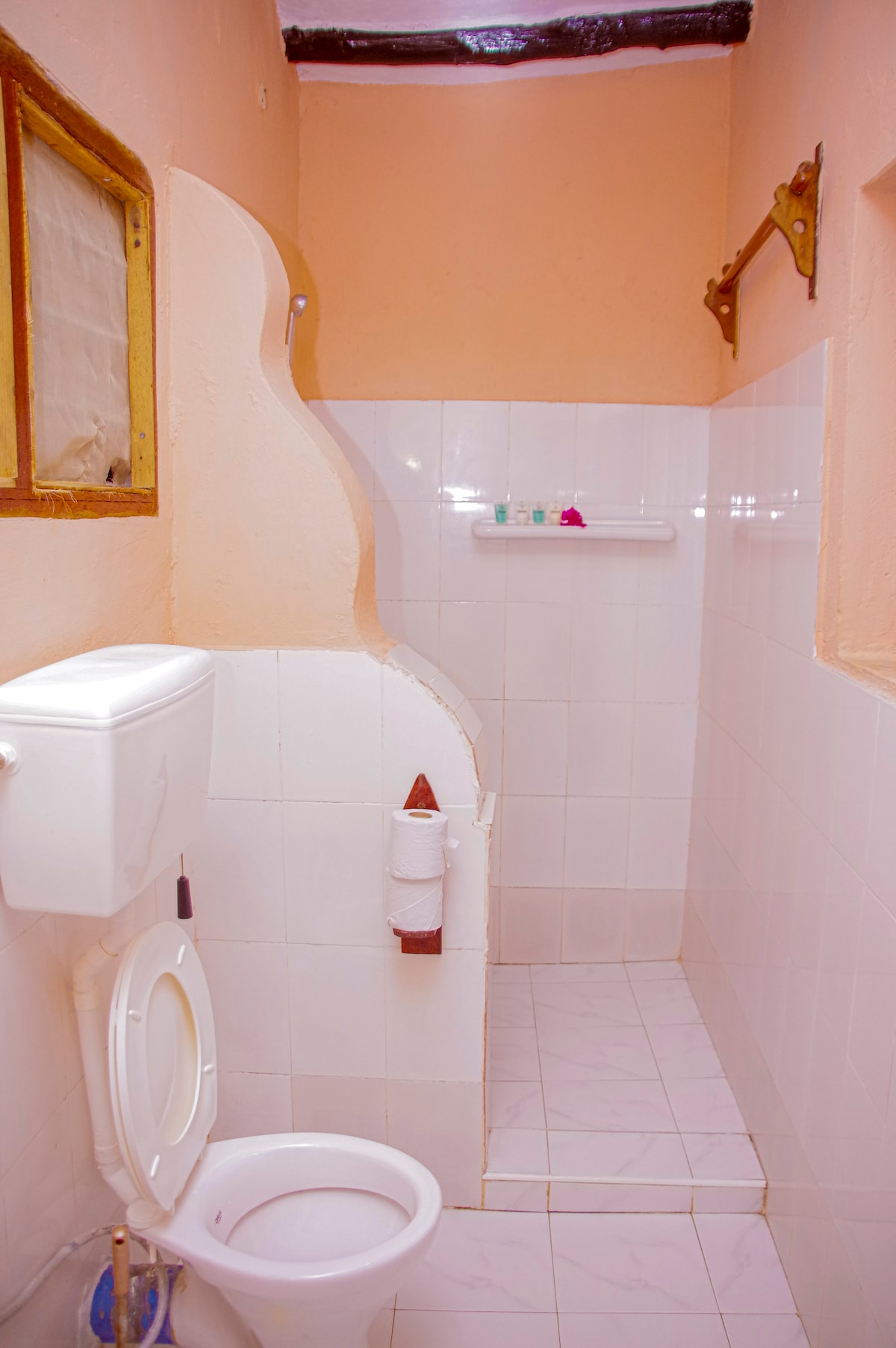 A clean bathroom space is characterized by white tiled walls and floor. A toilet is positioned next to a small shower area with a clear showerhead. A shelf holds toiletries, contributing to a functional and tidy environment.