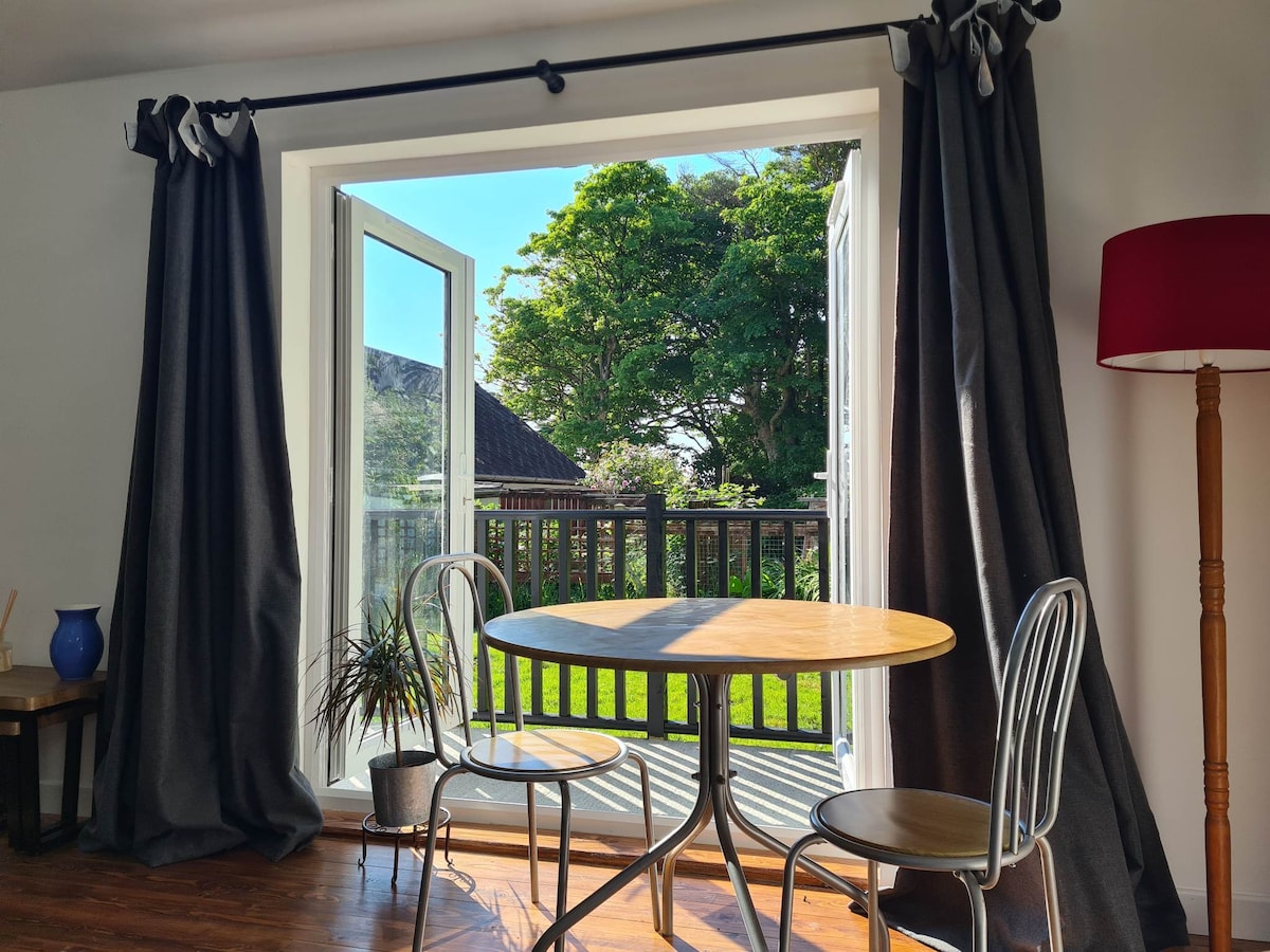 A bright dining area features a round wooden table complemented by two metal chairs. Open double doors lead to a balcony with a view of lush greenery. Large windows allow natural light to fill the space, while floor-to-ceiling curtains frame the scene.