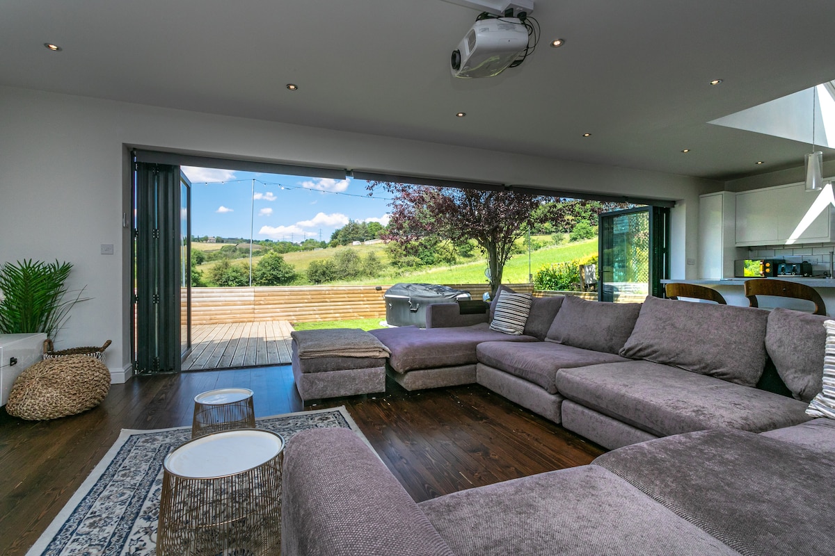 A spacious living area features a large sectional sofa, with natural light streaming in through expansive folding glass doors. A hot tub is visible on the deck outside, while lush green hills serve as a backdrop. Decorative side tables and a plant accent the room.
