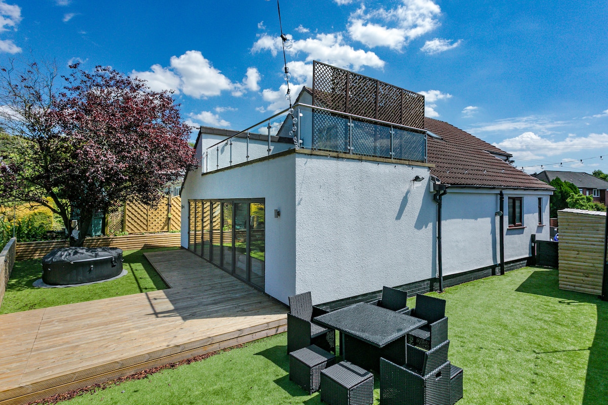 An expansive outdoor area is presented, featuring a well-maintained lawn and a wooden deck. A hot tub is situated nearby, and a patio set with multiple chairs is arranged for outdoor dining. Large glass doors provide access to the interior, with a rooftop terrace above.