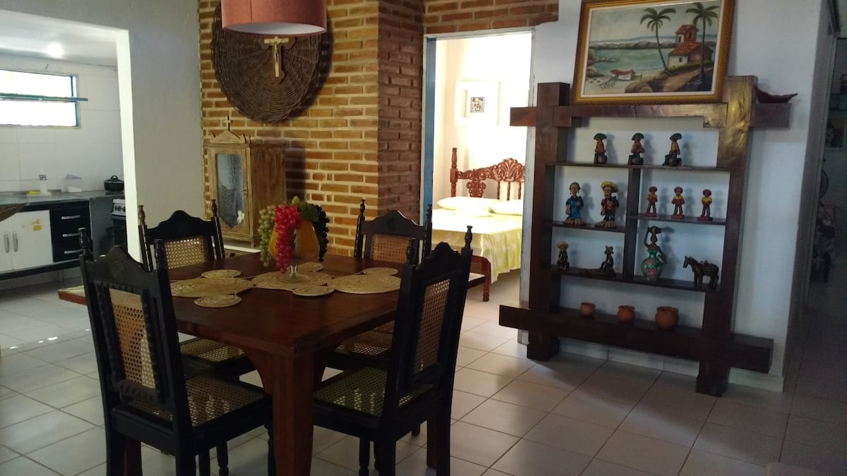 A dining area features a wooden table surrounded by six woven chairs. A decorative centerpiece of fruit is on the table. The room showcases brick walls, wooden accents, and a side display shelf filled with figurines, enhancing the warm ambiance of the space.