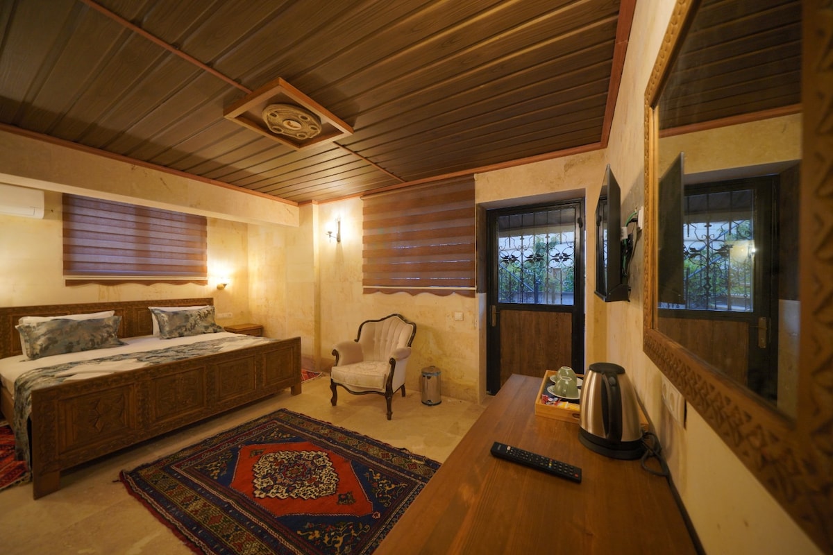 A tastefully decorated bedroom features a spacious bed crafted from dark wood, accompanied by a comfortable armchair. Natural light filters through patterned shades on the windows, revealing a richly colored area rug on the stone floor. A small table holds a kettle and remote control.