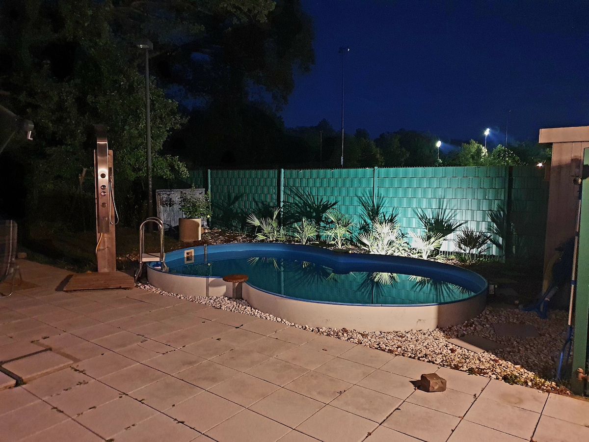 A tranquil swimming pool is set amidst a serene outdoor space, surrounded by lush greenery. Soft lighting illuminates the area, reflecting off the water. Paved stones create a sleek pathway, enhancing the relaxing environment, while a privacy fence provides seclusion.