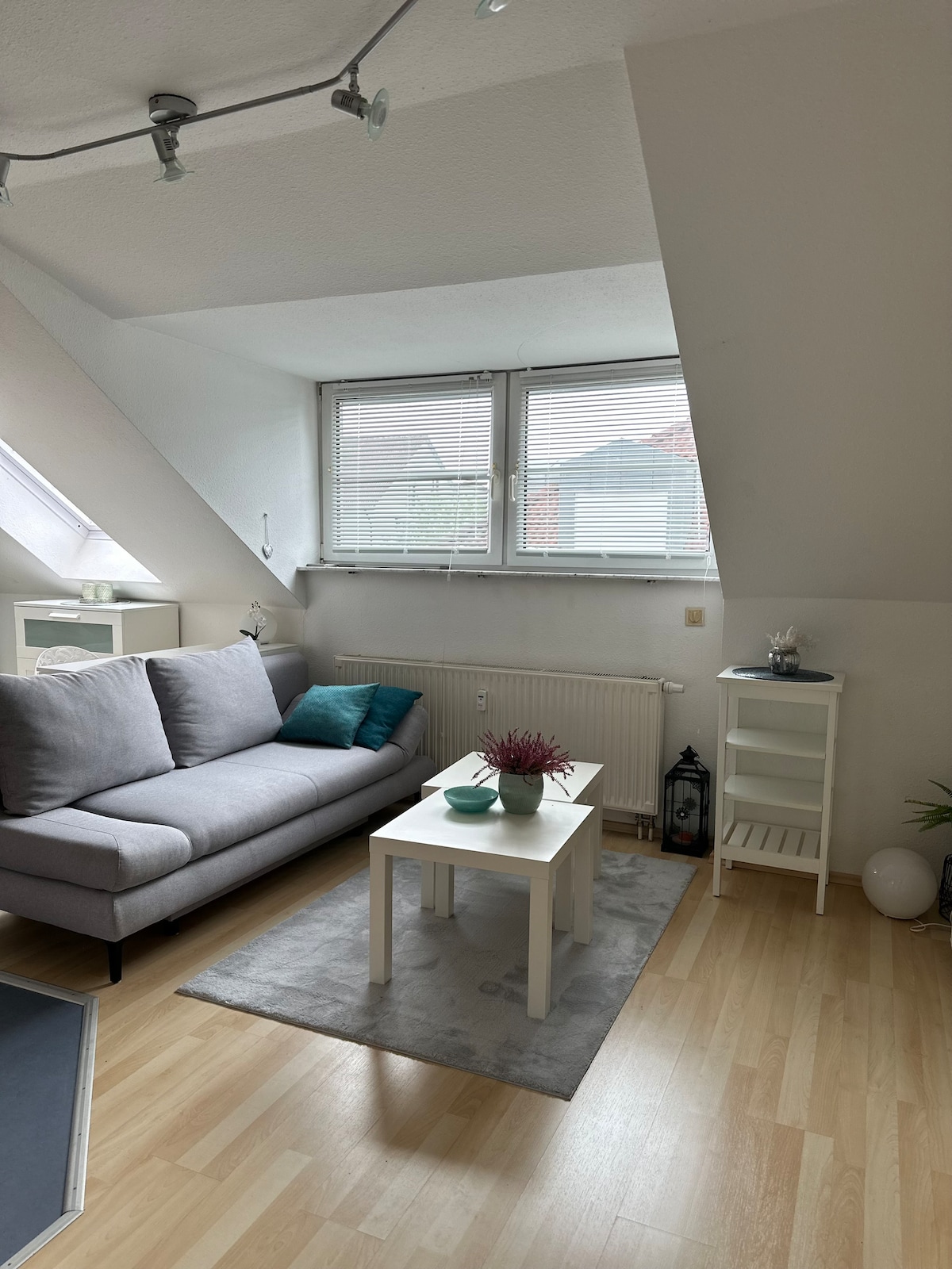 A cozy living area features a light gray sofa positioned beside a small white table. A decorative plant is placed on the table, complemented by additional plants nearby. Two large windows allow natural light to enter, with light-colored walls and a softly textured rug enhancing the space.