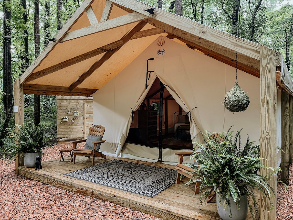A glamping tent is nestled under trees, featuring a wooden porch with two Adirondack chairs flanking a patterned area rug. The front opening of the tent provides a view into an inviting interior. Potted ferns add a touch of greenery to the entrance.