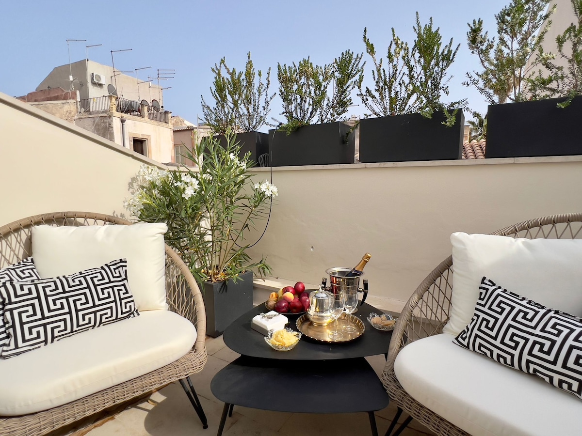 An outdoor terrace features two woven lounge chairs with plush cushions, positioned around a low table. On the table, a silver ice bucket holds a chilled bottle, accompanied by fruit and snacks. Potted greenery adds a touch of nature to the serene space.