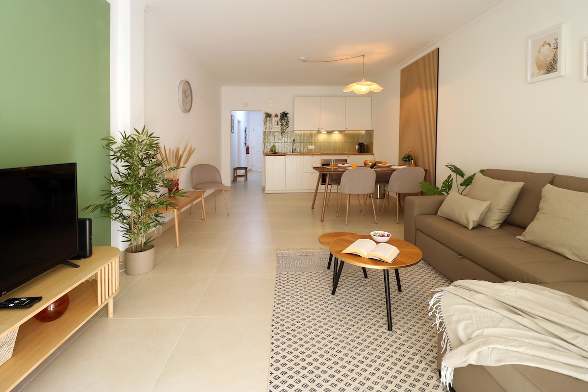 A spacious living area is presented with a comfortable sofa, coffee table, and a television. Decorative plants are positioned around the room, while a dining table with chairs is visible in the background. Natural light illuminates the modern design featuring neutral tones.