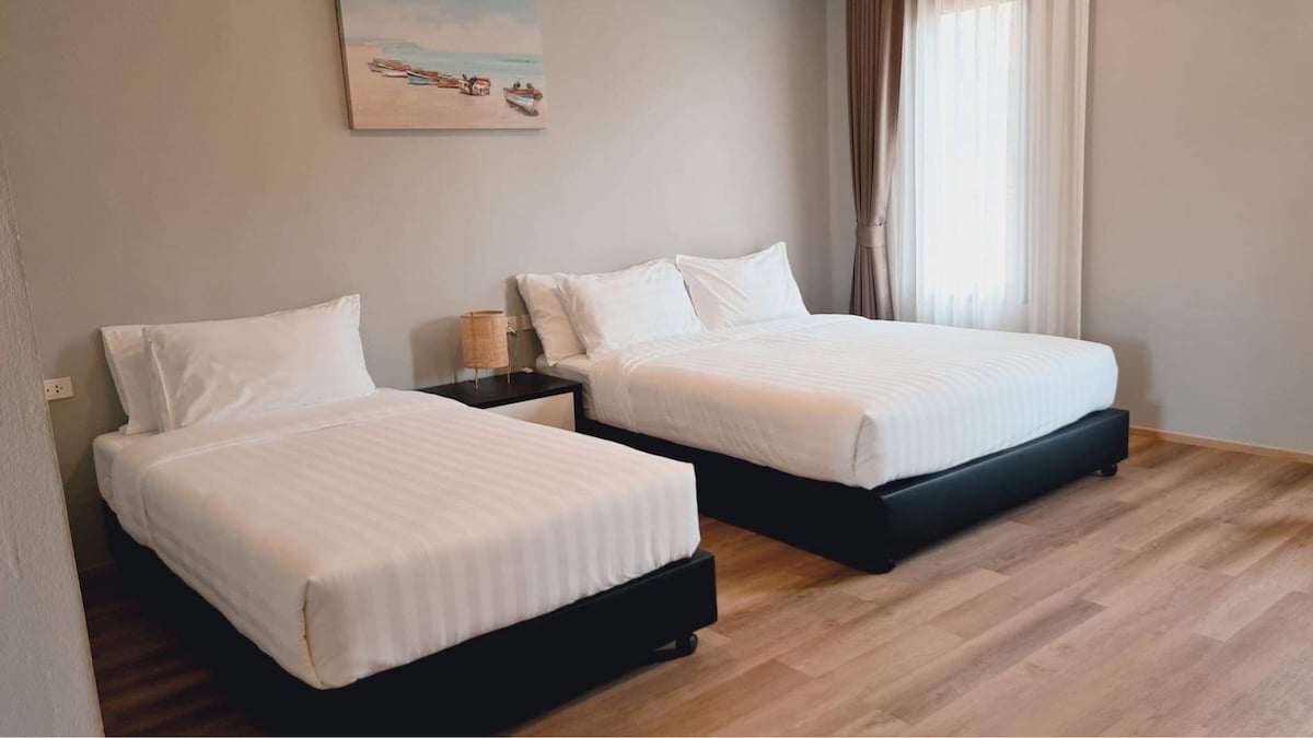 The sleeping area contains two spacious beds with crisp white linens, arranged against a neutral-colored wall. A piece of art depicting a beach scene adorns the wall above, while natural light filters through sheer white curtains of a nearby window.