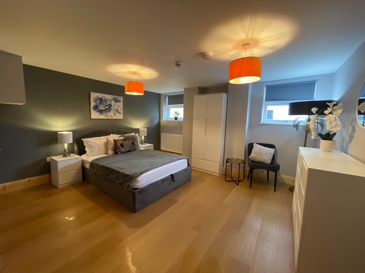 A spacious bedroom features a comfortable double bed dressed in white linens. Two bedside lamps provide soft lighting, and a wardrobe stands nearby. Natural light enters through two windows, with modern decor including a wall artwork and a chair accentuating the area.